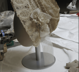 Side image of a cream lace cap embroidered with raised gold swirling motifs and scalloped edging, mounted on a display stand in a studio workspace.